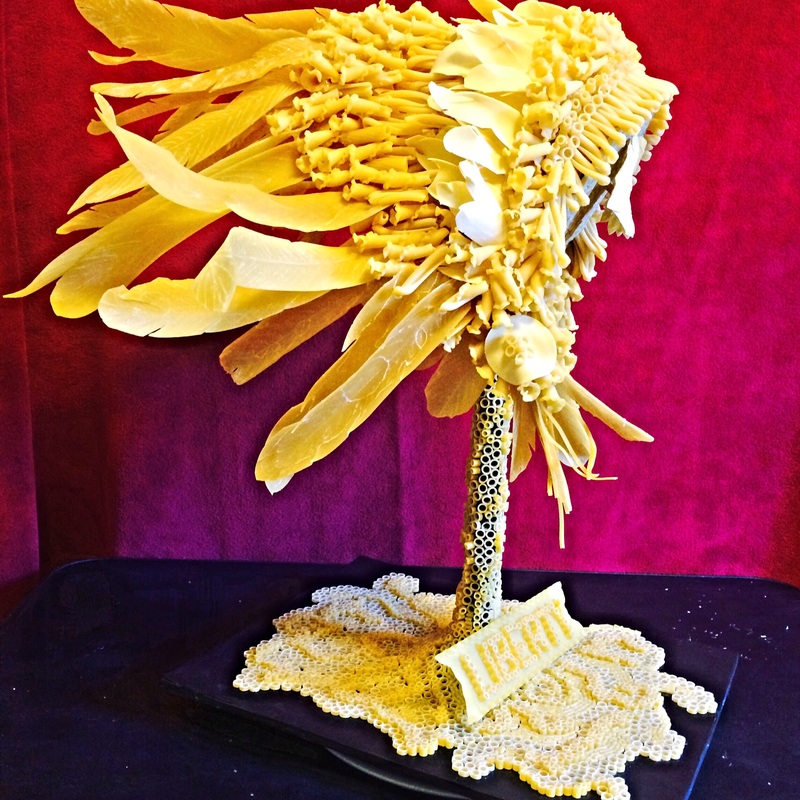 A sculpted palm tree crafted entirely from various types of yellow and white pasta stands on a black tray against a red and purple background.