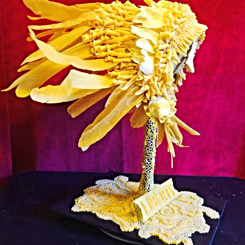 A sculpted palm tree crafted entirely from various types of yellow and white pasta stands on a black tray against a red and purple background.