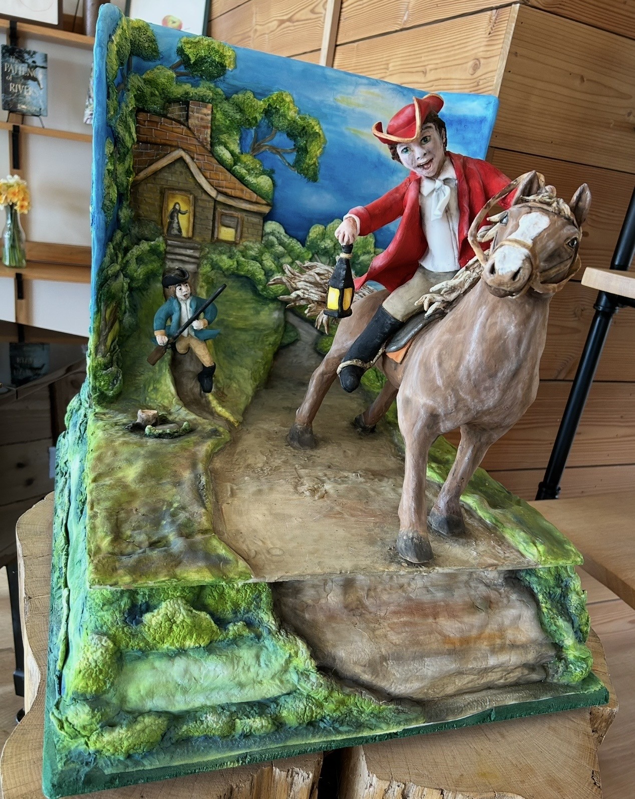 A dramatic historical diorama cake inspired by Paul Revere's midnight ride, featuring a young man in red tricorn hat and coat riding a brown horse with white blaze holding a lantern, smaller figure in blue colonial attire running with musket, rustic brick house with glowing windows, layered terrain of grassy slopes and rocky outcrops.