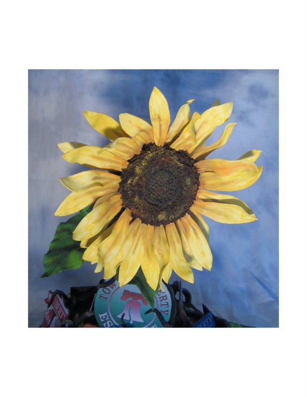 A large sunflower with yellow petals and a dark brown center stands against a blue sky background, positioned above a cluster of colorful party hats.