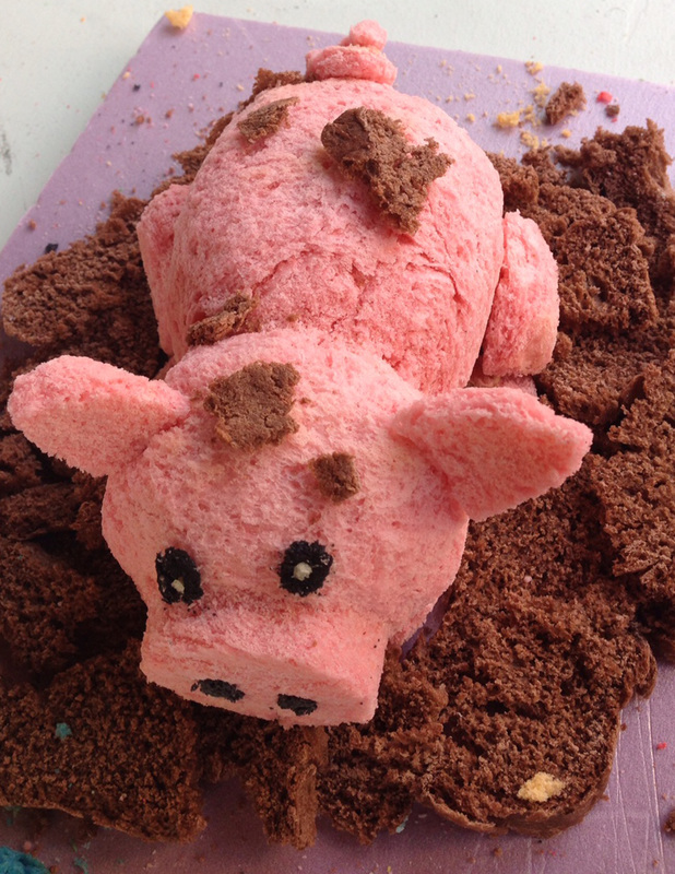 A pink sugar-crafted sculpture shaped like a cow with brown spots and black eyes rests in a pile of crumbled brown sugar or cookie sand on a purple surface.