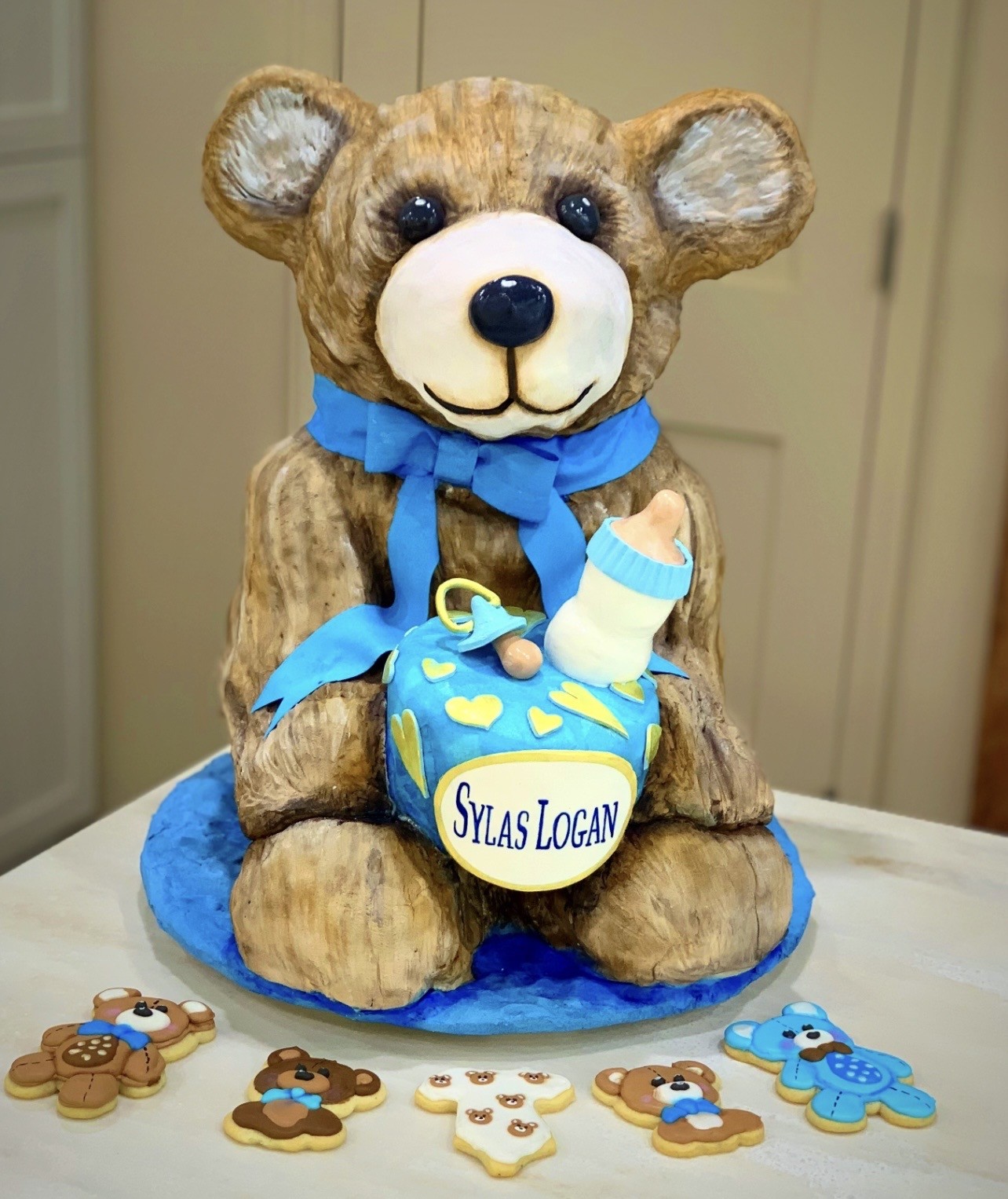 A sculpted cake designed as a seated brown teddy bear with textured icing fur, glossy black eyes, and a blue ribbon bow tie, holding a small round cake decorated in blue and yellow with heart motifs, a fondant baby bottle, pacifier, and a personalized plaque reading "Sylas Logan," surrounded by five teddy bear and onesie-shaped cookies.