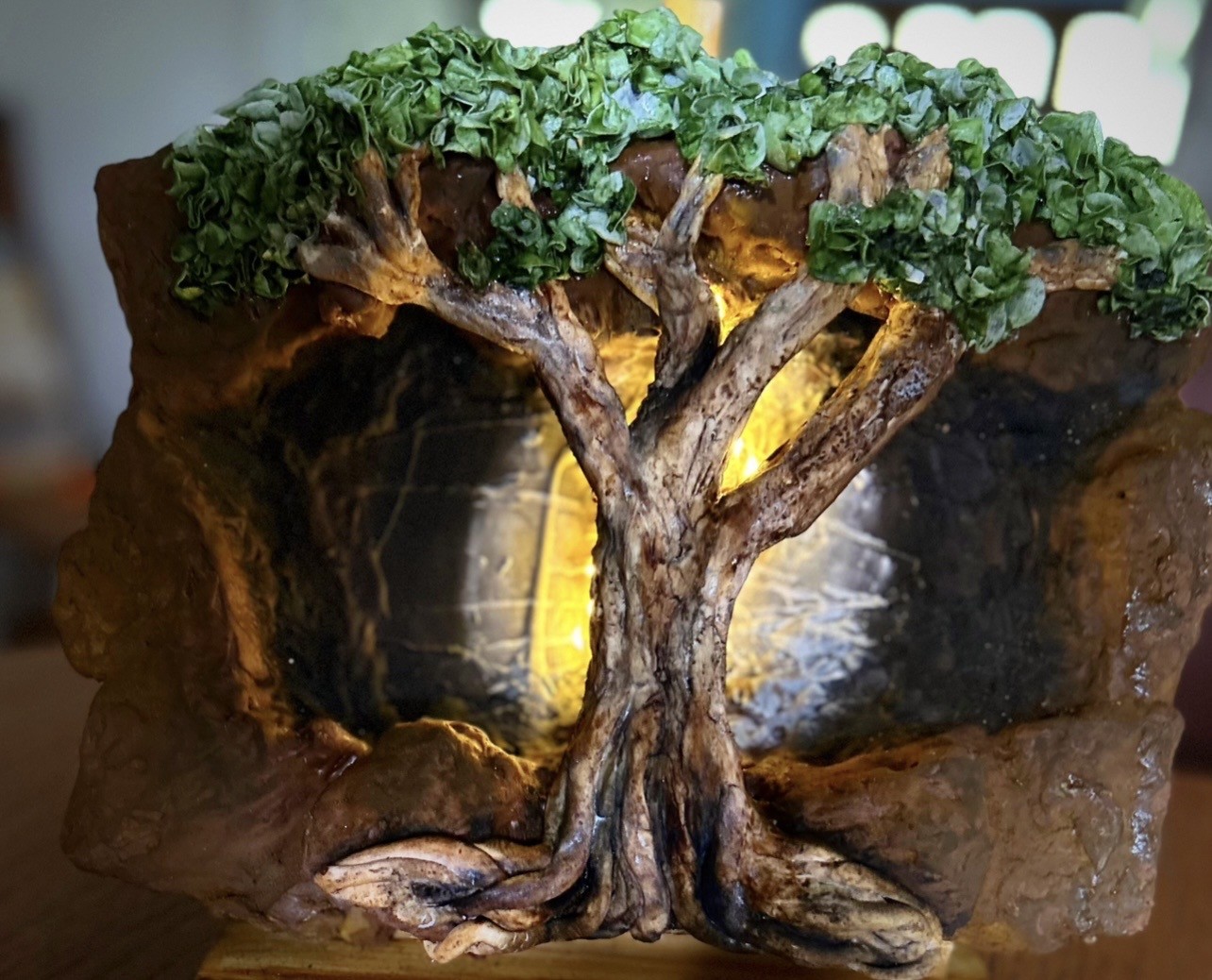 A sculpted cake diorama of a gnarled ancient tree emerging from a hollowed-out stone base, rich earthy brown textured bark, dense ruffled green foliage canopy, warm golden light emanating from within the cave-like hollow behind the tree, rough-hewn rock structure in ochre, sienna, and charcoal.