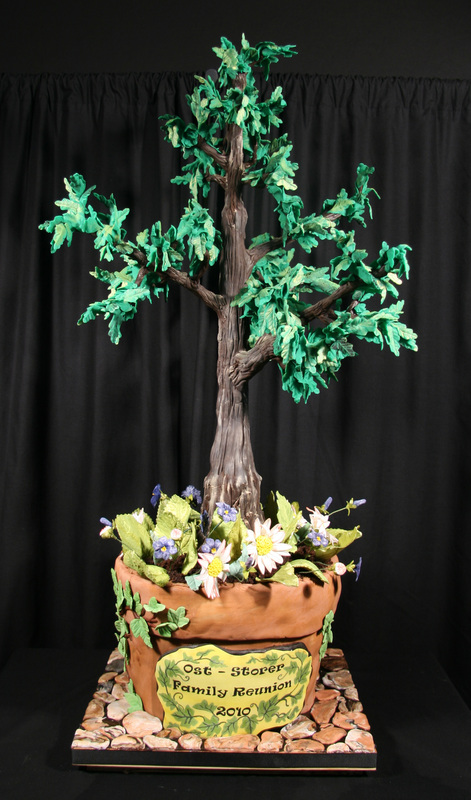 An elaborate cake designed to look like a potted tree with green leaves and a brown trunk, surrounded by fondant flowers and a plaque reading "Ost - Storer Family Reunion 2010."