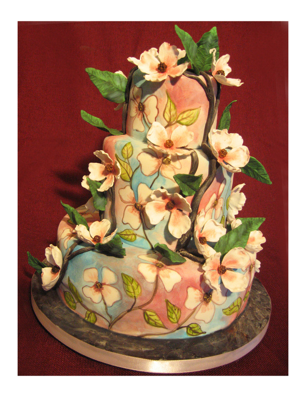A three-tiered cake decorated with pastel pink and blue fondant, featuring hand-painted floral patterns and adorned with sculpted white flowers and green leaves.