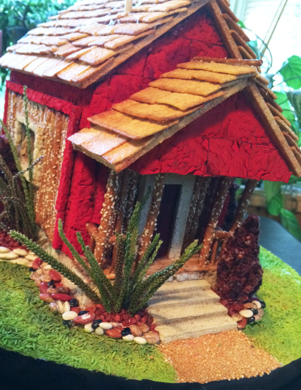An elaborate gingerbread house features red icing walls, a roof made of rectangular crackers, and a landscaped base with green dyed rice, beans, and a succulent plant.