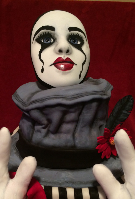 A sculpted cake designed to look like a mime, featuring a white face with black tear-like makeup, red lips, and a grey ruffled collar adorned with a red flower and black feather.