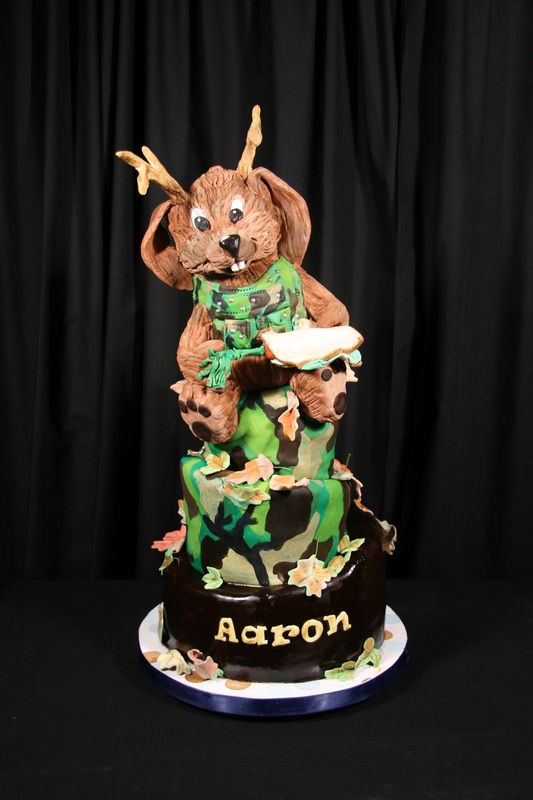A multi-tiered camouflage-patterned cake features a sculpted brown bunny wearing antlers and a camo vest, with the name "Aaron" written in gold icing on the chocolate base.