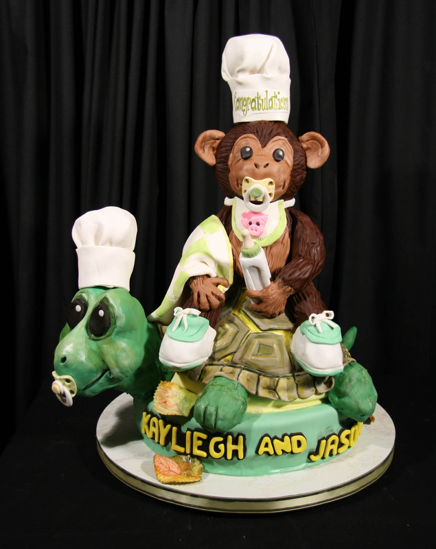An elaborate sculpted cake features a baby monkey wearing a chef's hat and bib riding on the back of a green turtle, also wearing a chef's hat, with the names "KAYLIEGH AND JASON" written in yellow letters on the base.