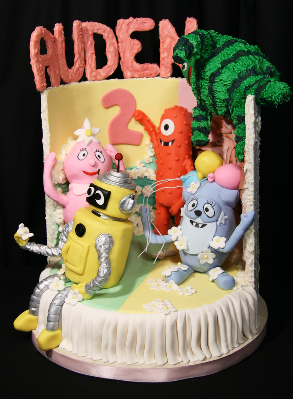 A colorful birthday cake features fondant characters from the children's show "In the Night Garden," including Igglepiggle, Upsy Daisy, Makka Pakka, and Wotting, along with a large number "2" and the name "AUDEN" in pink letters on top.