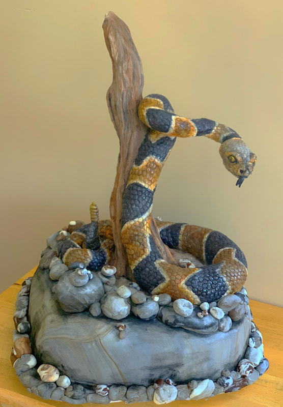A sculpted cake features a detailed, three-dimensional snake with black and orange bands coiling around a vertical piece of wood, set upon a rocky, stone-textured base.