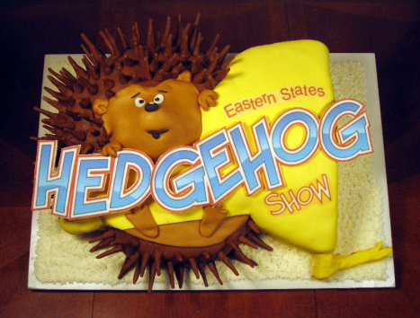 A square sheet cake features a 3D fondant hedgehog character wearing a yellow blanket, accompanied by large blue and red lettering that reads "Eastern States HEDGEHOG SHOW."