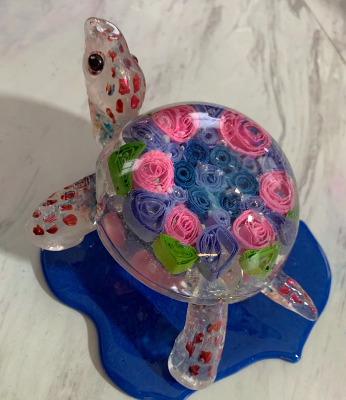 A clear sugar glass sea turtle figurine with a shell filled with pink, blue, and green paper-like sugar quilling roses, featuring red speckled flippers and a black bead eye, resting on a blue sugar base.