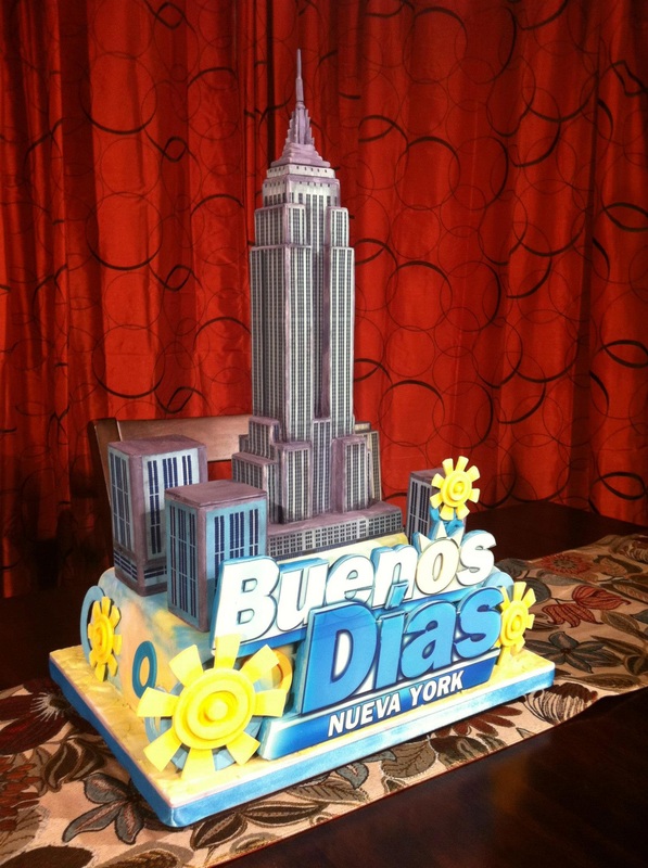 A fondant cake designed as a New York City skyline features a tall Empire State Building replica, smaller skyscrapers, and the words "Buenos Dias NUEVA YORK" in blue and white lettering, all decorated with yellow sun motifs and set against a red curtain backdrop.