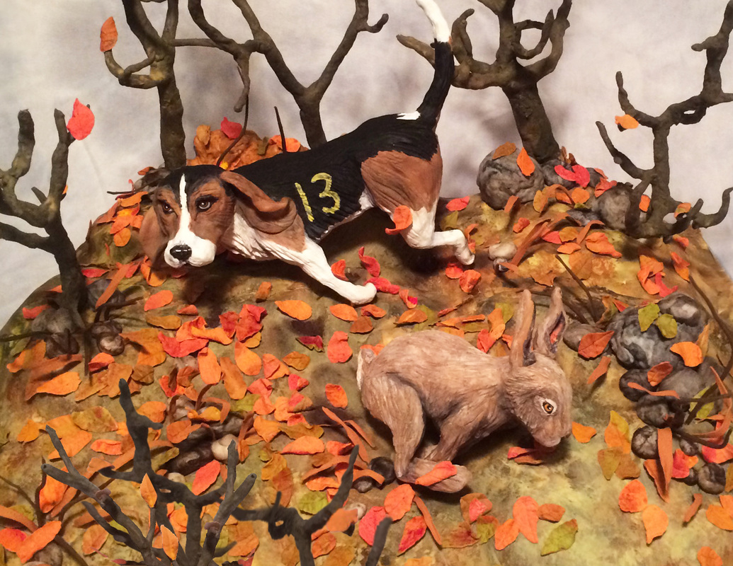 A diorama depicts a tricolor hound wearing a black vest with the number 13 in pursuit of a brown hare through an autumnal forest floor scattered with orange leaves and bare trees.