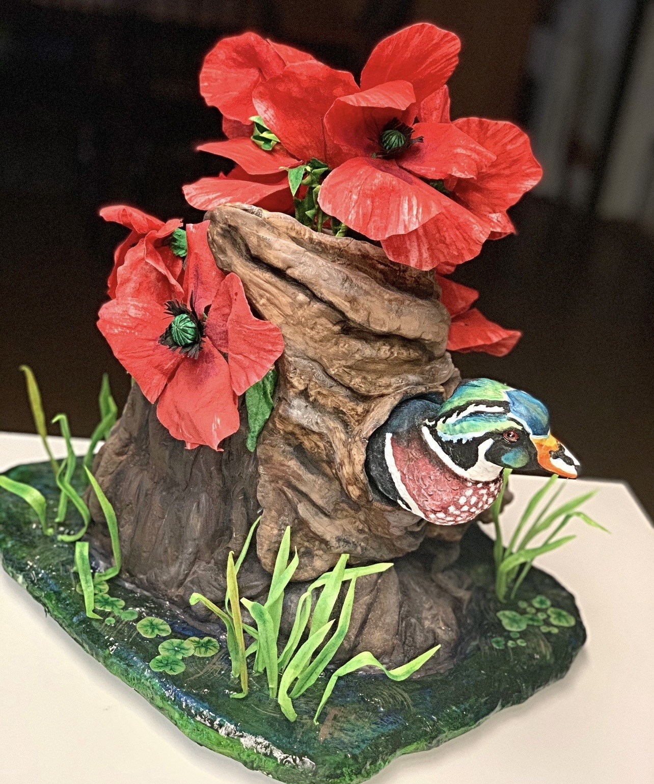 A sculpted cake diorama featuring a textured tree-stump-like structure in earthy browns and grays, vibrant red poppies with visible veins emerging from the top, hollow containing head of a male wood duck with iridescent green, blue, white plumage and striking orange beak, surrounded by lush green landscape on glossy dark green surface.