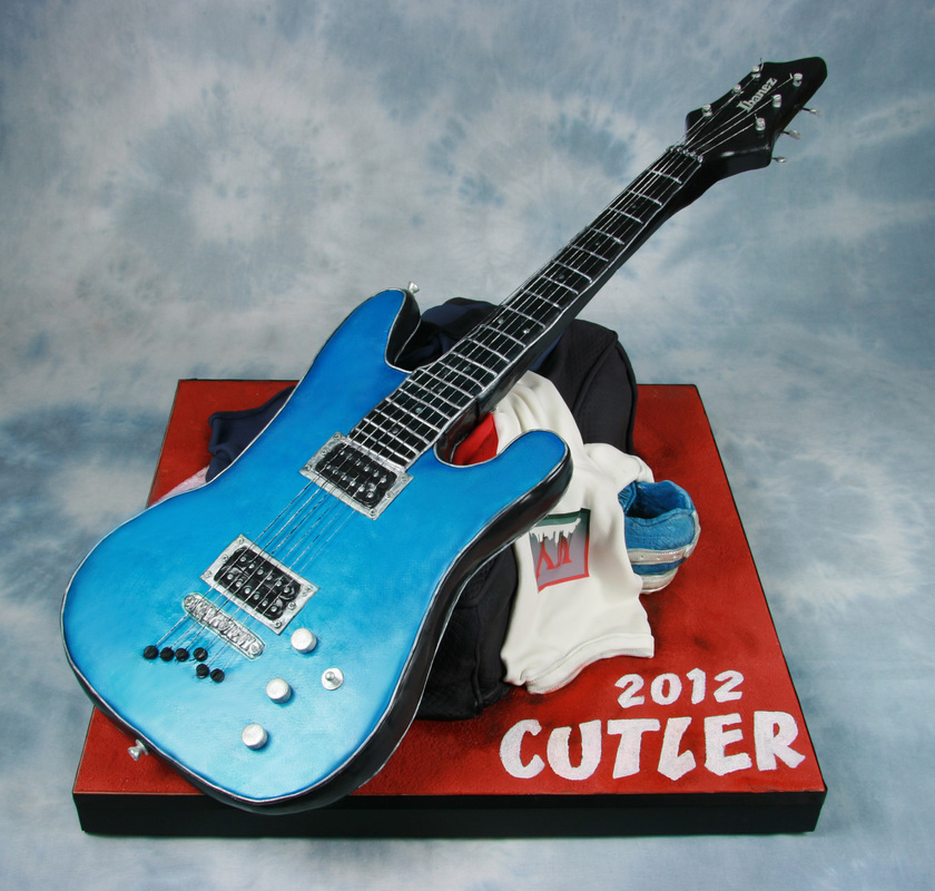 A blue electric guitar rests on a red display board labeled "2012 CUTLER," accompanied by a white t-shirt and a blue shoe.