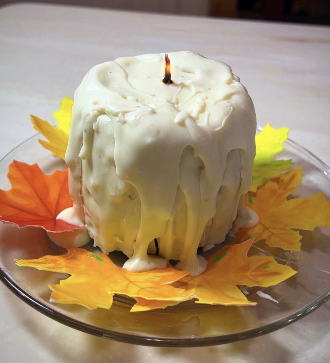 A whimsical cake designed to resemble a melting white candle with thick off-white icing dripping down its sides like molten wax, a thin dark wick protruding from the top, surrounded by artificial autumn leaves in vibrant orange, yellow, and green arranged in a circular pattern on a clear glass plate.
