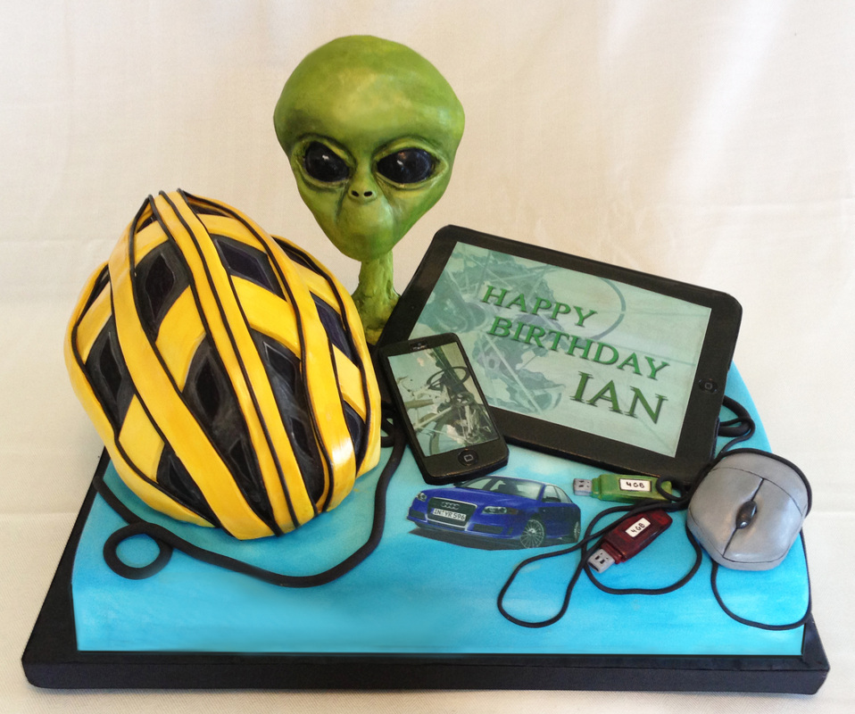 A blue birthday cake decorated with fondant figures of a yellow bicycle helmet, a green alien head, a tablet displaying "Happy Birthday Ian," a smartphone, a blue car, USB drives, and a computer mouse.