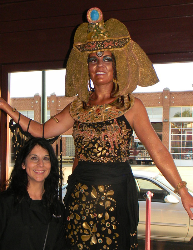 A smiling woman with long dark hair stands next to a life-sized statue of Cleopatra wearing an elaborate golden headdress and black dress with gold embellishments.