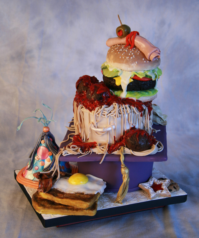 An elaborate, multi-tiered cake designed to resemble a grotesque breakfast scene features a giant hamburger with dripping condiments perched atop a bucket overflowing with spaghetti and red sauce, accompanied by fondant bacon, a fried egg, and a party hat on the base.