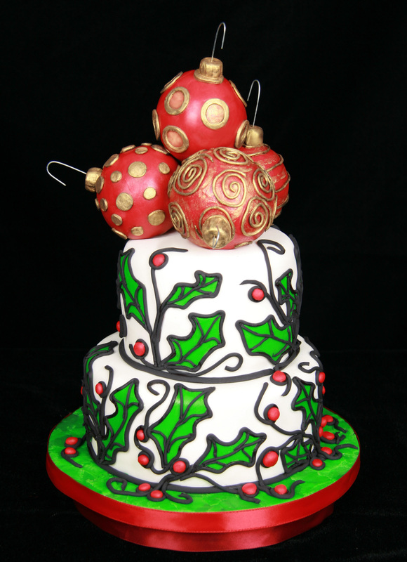 A two-tiered white Christmas cake decorated with green holly leaves and red berries, topped with three large red and gold fondant ornaments.