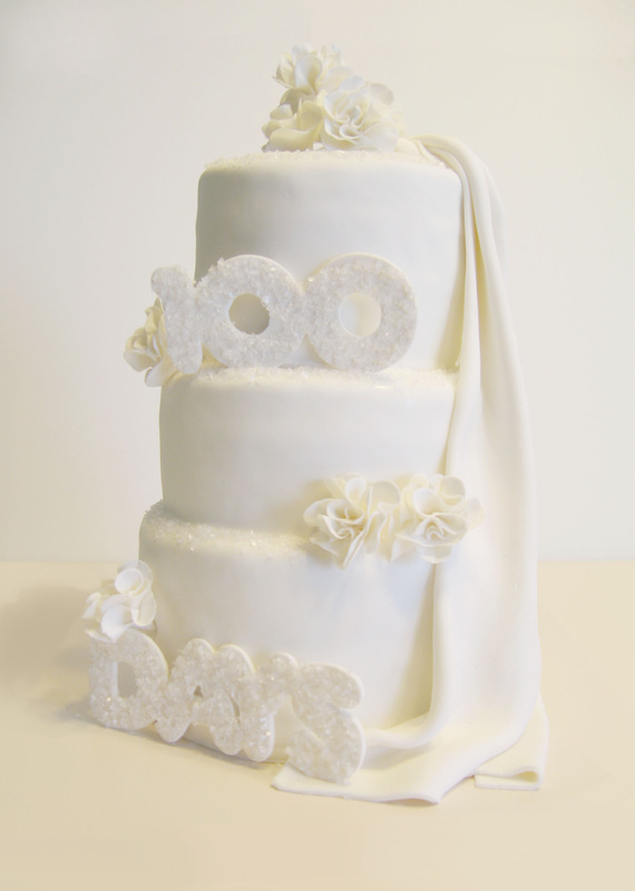 A three-tiered white fondant wedding cake adorned with sparkling "100" and "DAYS" lettering, white sugar flowers, and a draped fabric veil.