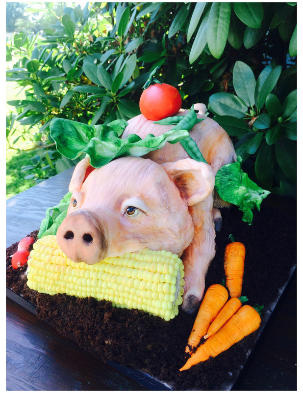 A hyper-realistic cake sculpted to resemble a pig chewing on a corn cob, surrounded by fondant carrots and lettuce on a dark base.