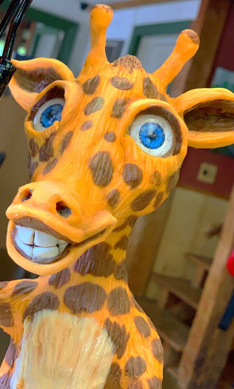 A close-up of a whimsical, hand-carved wooden giraffe sculpture featuring bright blue eyes, a wide toothy grin, and brown spots painted on its orange body.