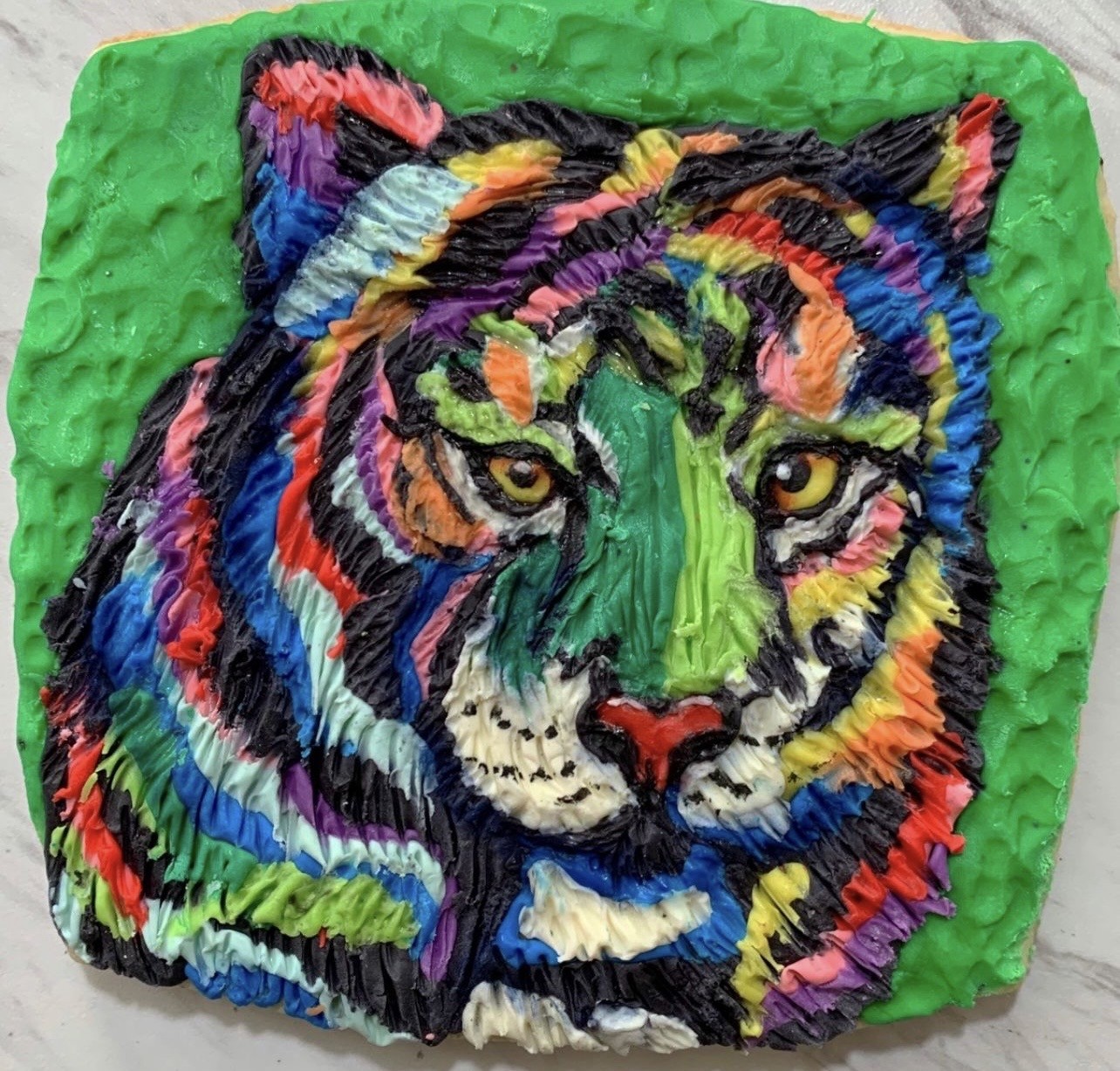 A vibrant textured cake decorated to resemble a psychedelic tiger's face against a bright green frosted background, with bold multicolored stripes in blues, purples, oranges, pinks, and greens blending into a tie-dye effect, thick piped frosting adding depth to the tiger's yellow eyes, nose, whiskers, and stripes.