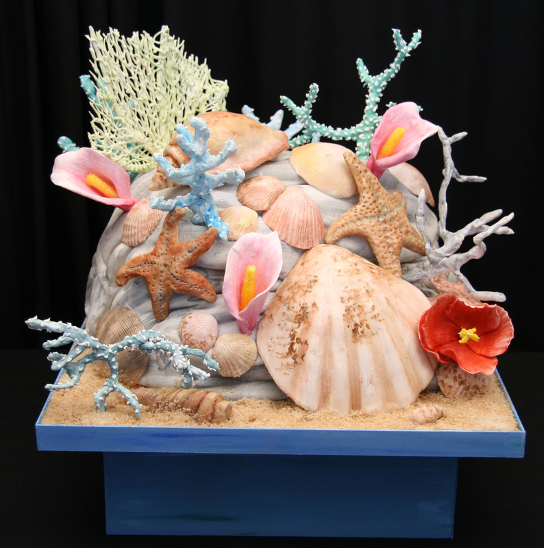 An elaborate cake decorated with fondant seashells, starfish, and coral sits on a blue base covered in edible sand.