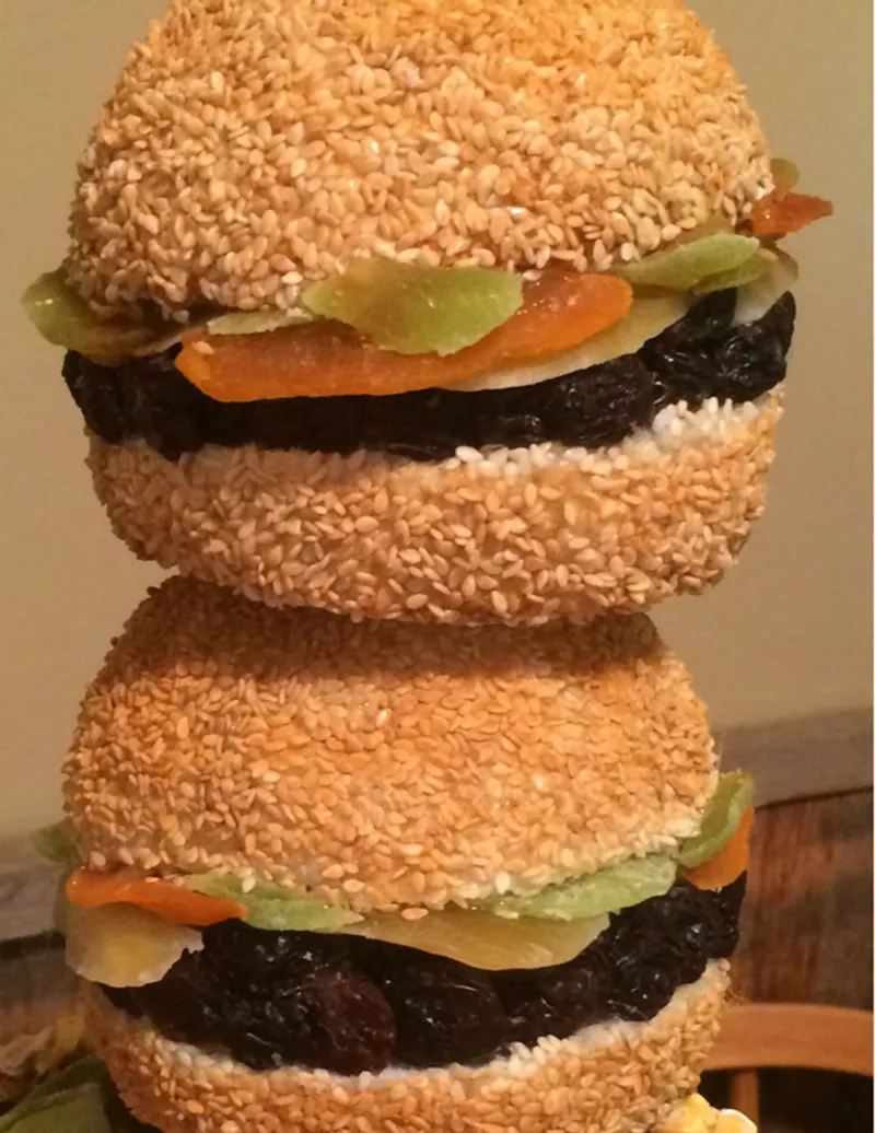 Two stacked sandwiches made with sesame seed buns, featuring dark patty-like fillings, sliced pickles, and orange peppers, displayed as a food art composition.