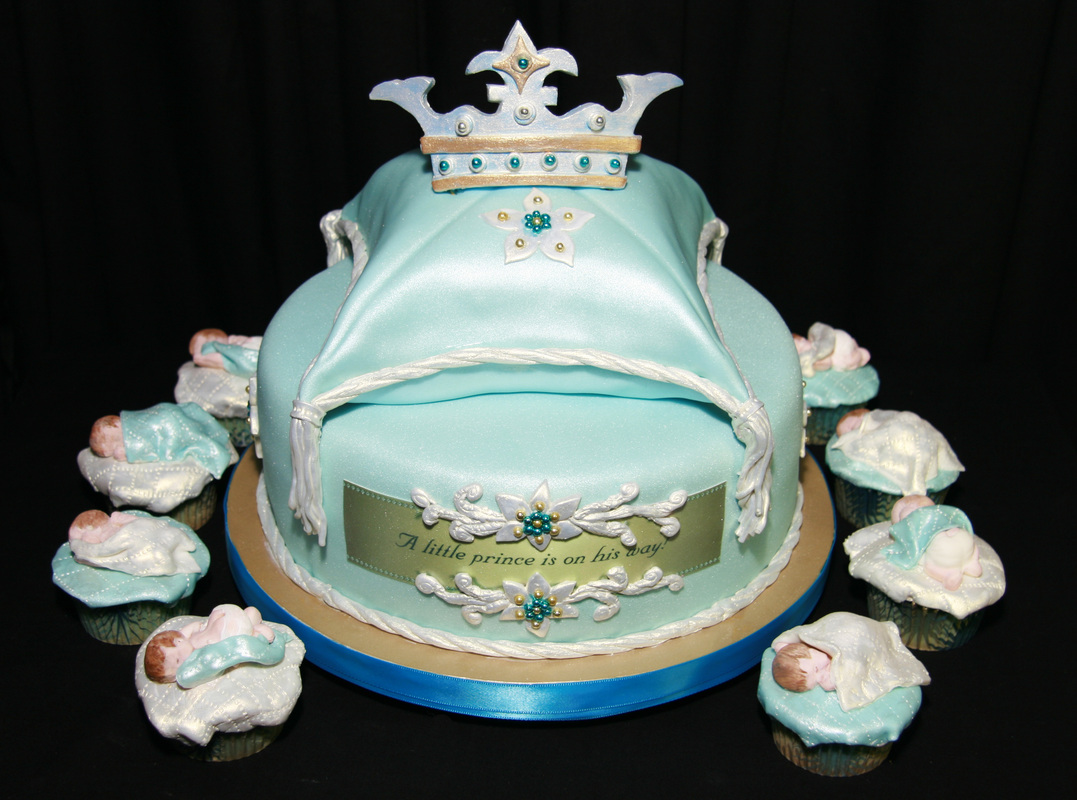 A light blue, crown-topped baby shower cake decorated with silver accents and the message "A little prince is on his way," surrounded by cupcakes topped with fondant sleeping babies.