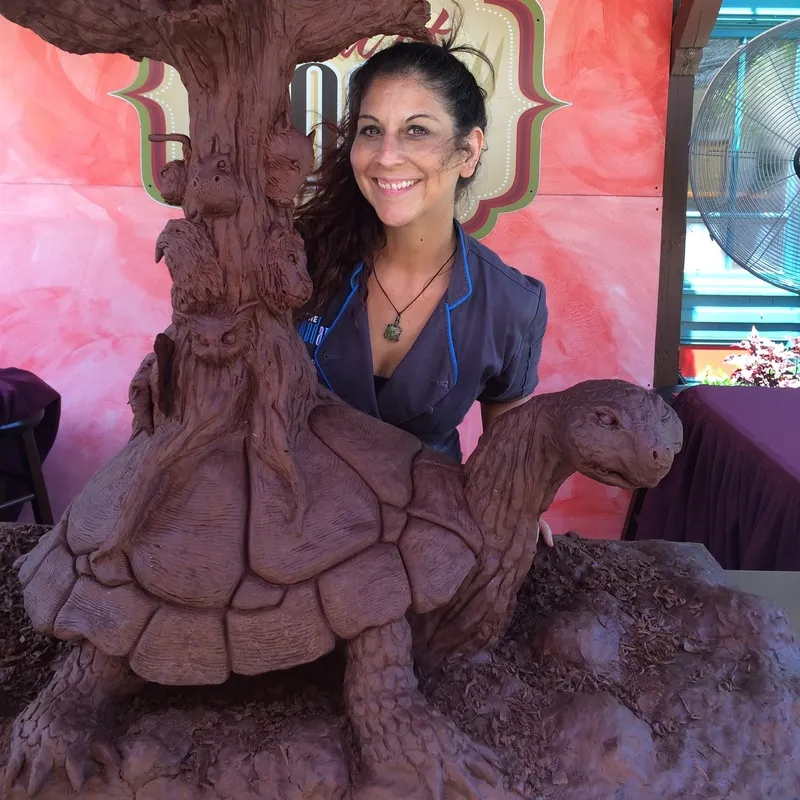 A smiling woman in a blue uniform stands beside a large, intricate chocolate sculpture of a tortoise carrying a tree with carved animals on its trunk.