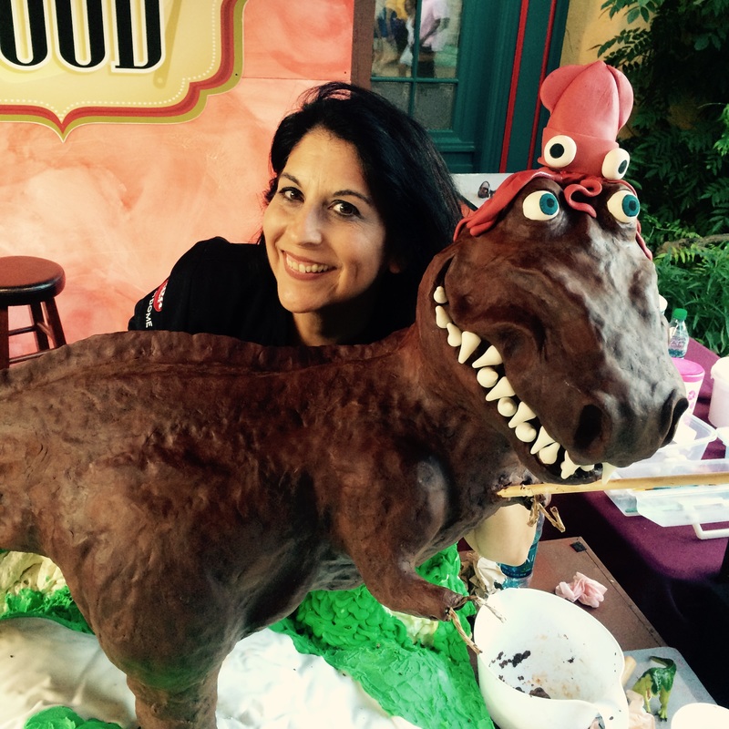 A smiling woman with dark hair poses behind a large, brown dinosaur cake decorated with white teeth and a red octopus topper, set against a backdrop featuring a sign with the word "FOOD."