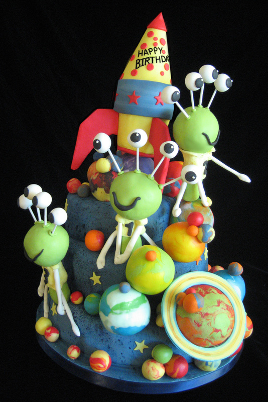 A space-themed birthday cake features a blue, star-speckled base adorned with colorful planets and three green, multi-eyed aliens, topped by a yellow and red rocket ship with "Happy Birthday" written on it.