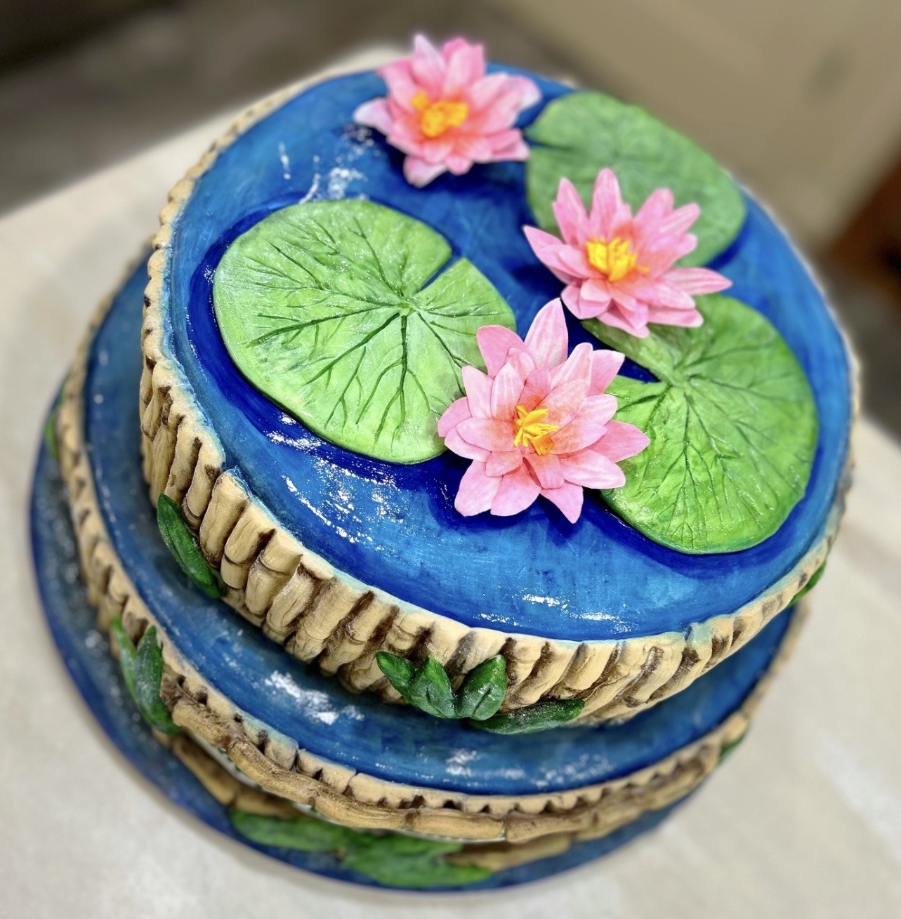 A two-tiered cake designed as a serene water lily pond with vibrant blue textured "water" surface mimicking ripples, three delicate pink water lilies with yellow centers crafted from fondant, large realistic green lily pads with radiating veins, sides adorned with woven basket-weave pattern in beige and brown tones.