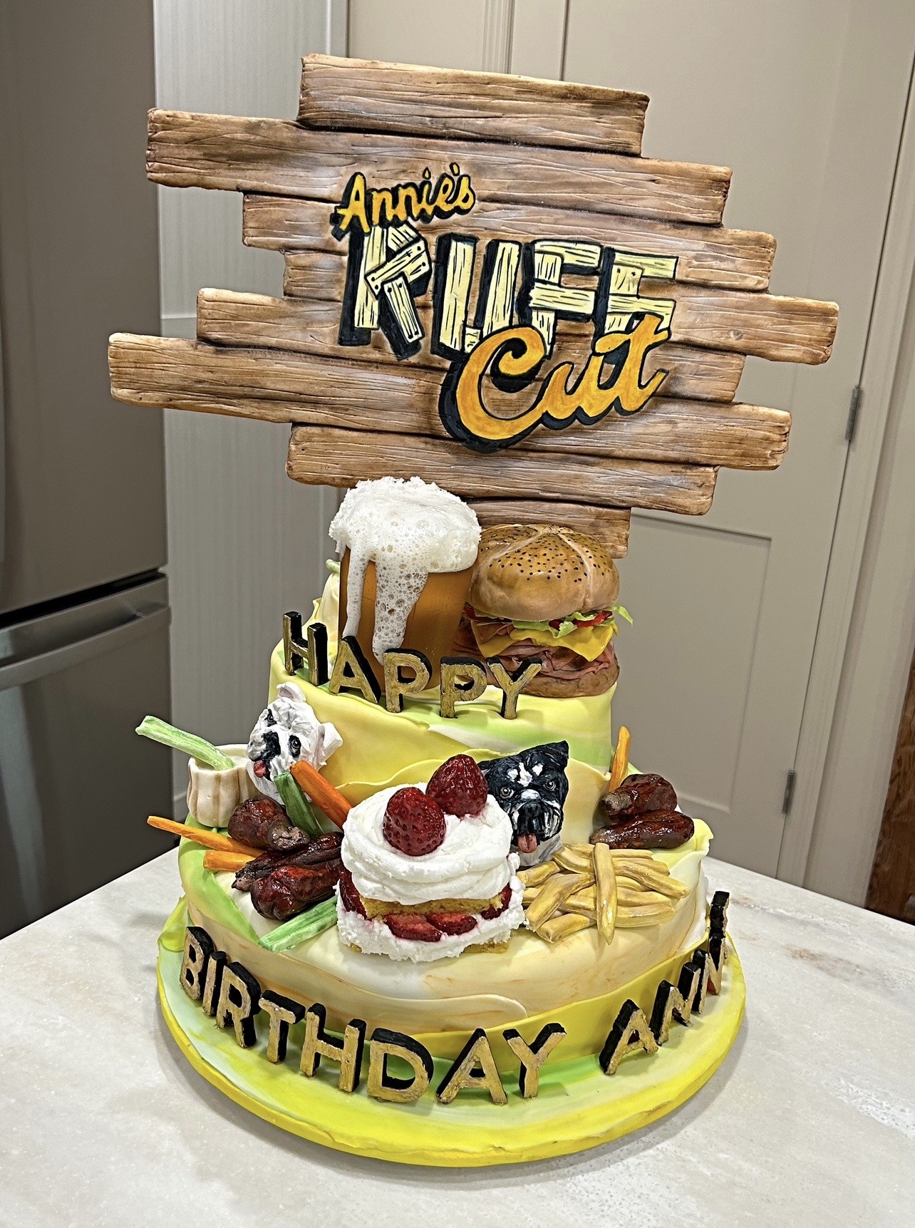 A rustic bar-and-grill themed birthday cake with fondant sign reading "Annie's Ruff Cut" in bold yellow and black, upper tier featuring realistic burger and frothy beer mug, lower tier with strawberry shortcake, french fries, glazed sausages, celery sticks, carrot sticks, small Boston Terrier figurine, and "BIRTHDAY ANN" in chunky black-and-gold block letters.