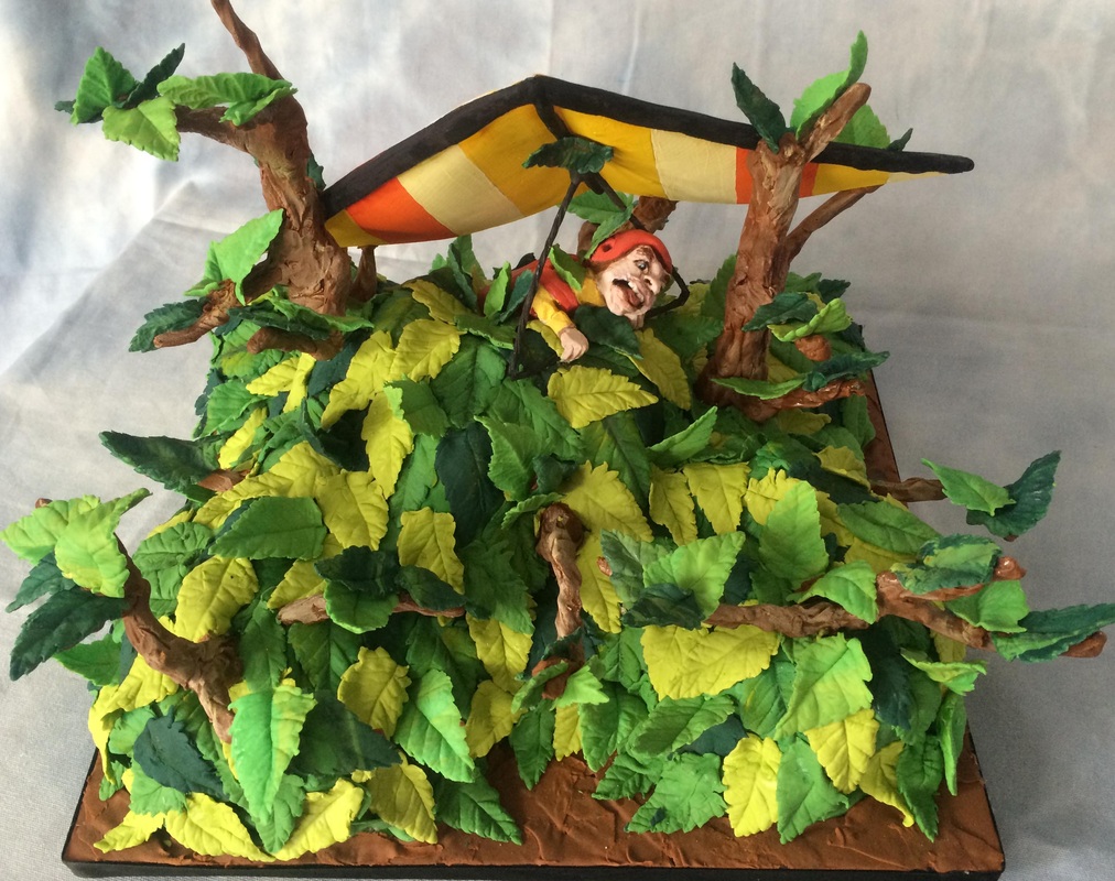 A sculpted cake diorama depicts a man in a red helmet and yellow shirt suspended in a striped hang glider between two trees, hovering over a dense canopy of green and yellow leaves.
