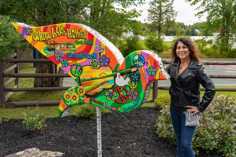 Kim Simons beside a painted dove sign for White Lake Homes with psychedelic art.