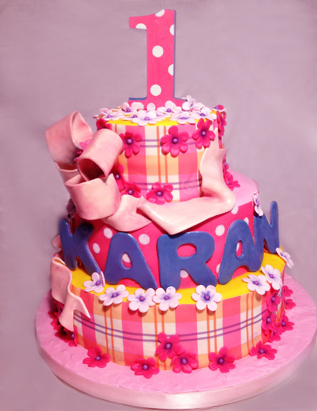 A two-tiered birthday cake decorated with pink and yellow plaid fondant, white polka dots, and pink flowers, featuring the name "KARAN" in blue letters on the bottom tier and a large pink number "1" topper on the upper tier.