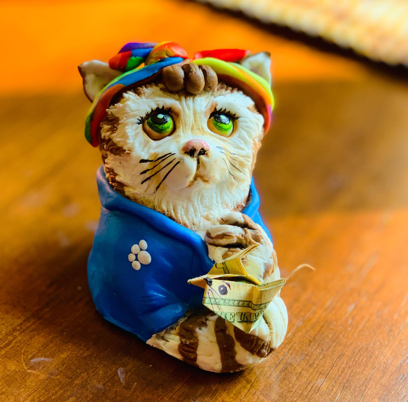 A small sugar figurine of a cat wearing a blue jacket and a colorful hat holds a tiny origami boat made from a dollar bill.