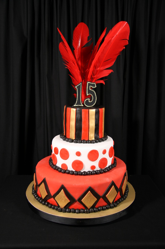 A three-tiered birthday cake in red, black, white, and gold features geometric patterns and polka dots, topped with red feathers and a number 15 topper.