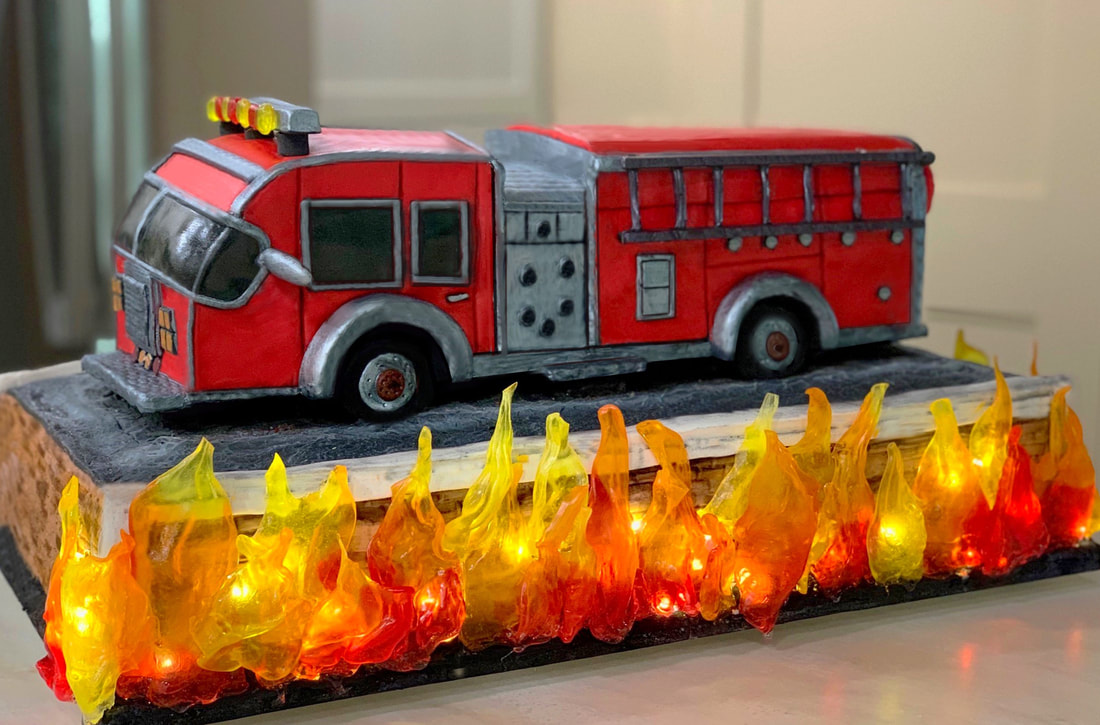 A detailed fire truck cake with red and silver icing sits on a black base, surrounded by translucent, flame-shaped candies illuminated from within by small lights.