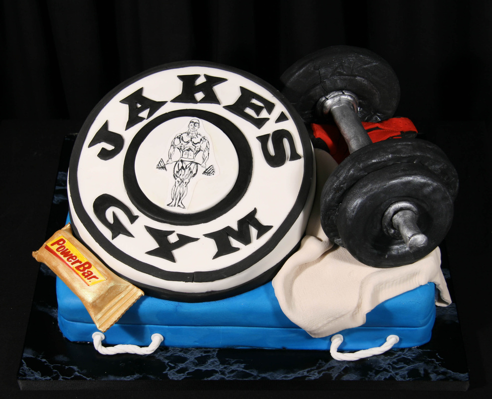 A gym-themed cake features a large fondant weight plate labeled "Jake's Gym" with a bodybuilder illustration, alongside a sculpted dumbbell, a Power Bar, and a white towel, all resting on a blue rectangular base.