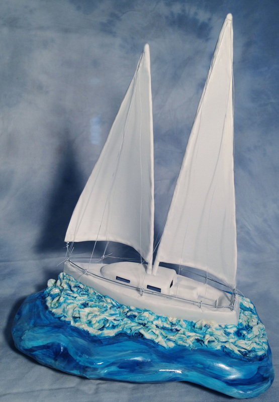 A sculpted cake of a white sailboat with two masts sits atop a sculpted blue and white base designed to resemble ocean waves.