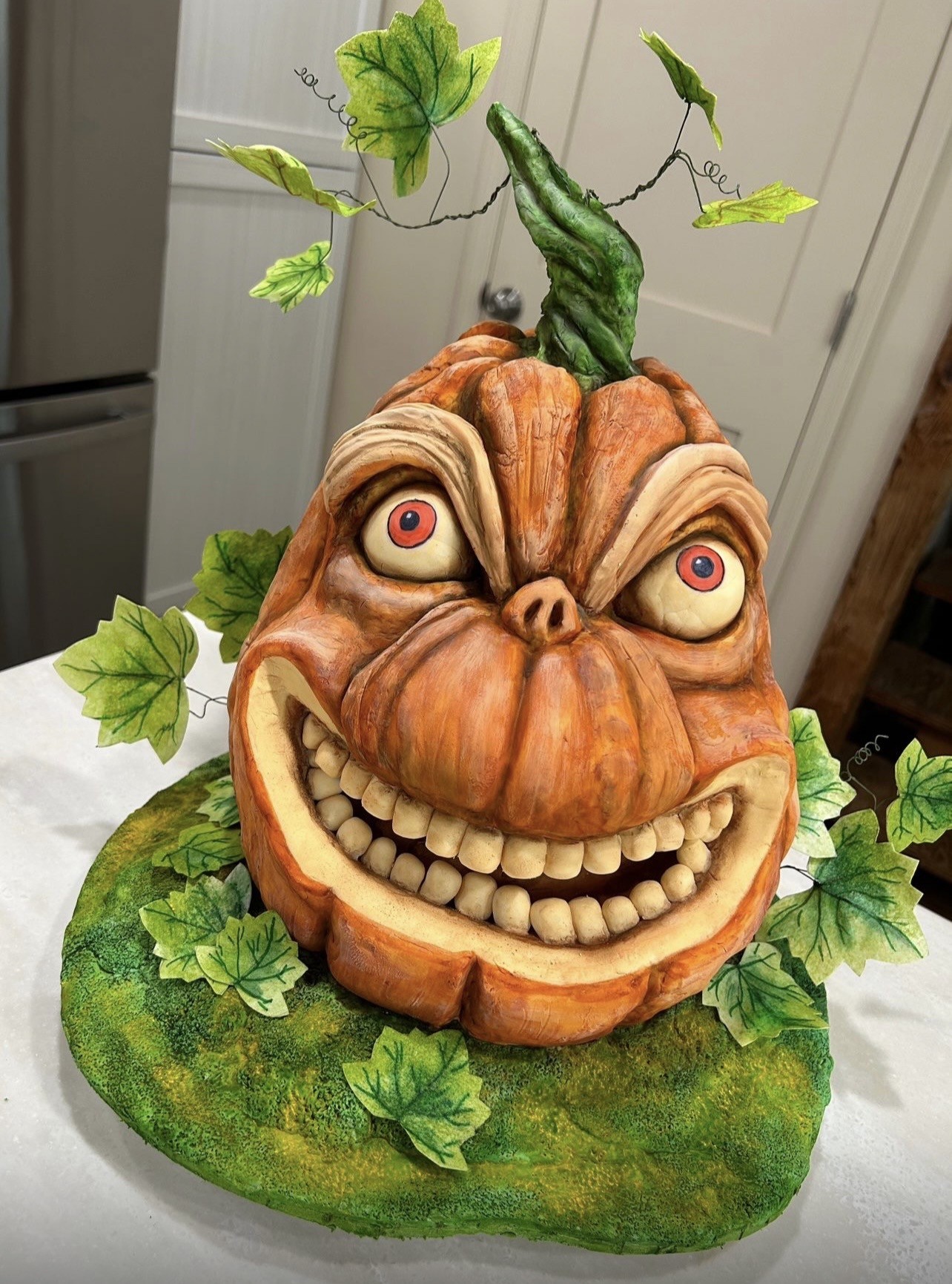 A sculpted cake designed as a grotesque anthropomorphic pumpkin head with bulging wide-set red-eyed eyes, broad toothy grin revealing uneven off-white fondant teeth, deeply furrowed brow ridges, rich textured orange and brown skin with realistic creases, twisted green stem, surrounded by hand-painted green ivy leaves on mossy-green platform.