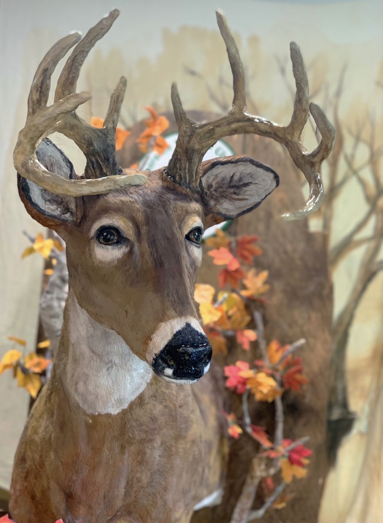 A sculpted cake of a white-tailed deer buck in hyper-realistic style with large soulful dark eyes, glossy black nose, impressive asymmetrical textured antlers in beige and gray, rich brown fur, prominent white patch on throat and chest, set against autumnal foliage backdrop in warm orange, red, and yellow.