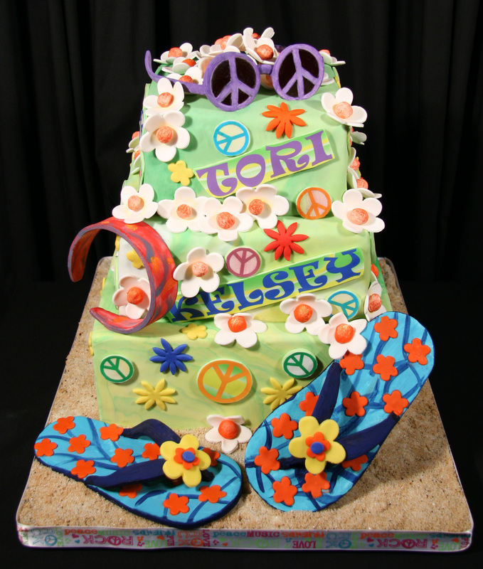 A three-tiered light green fondant cake decorated with peace signs, flowers, and the name "Tori Kelsey," accompanied by large fondant flip-flops and a crescent moon on a sandy base.