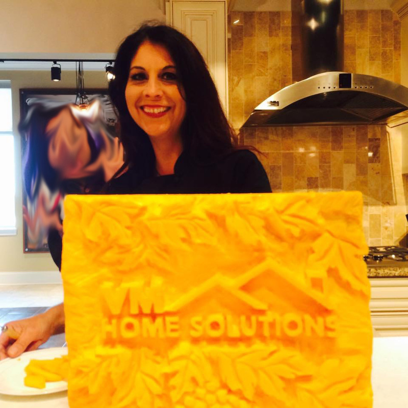A smiling woman with dark hair stands behind a large, intricately carved yellow block featuring the logo "VM HOME SOLUTIONS" and decorative leaf patterns.